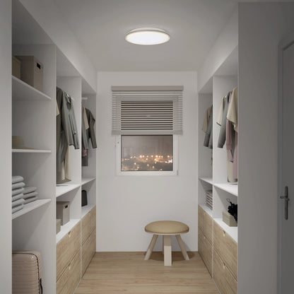 A modern walk-in closet features shelves, drawers, and hanging space on both sides, neat storage, a round stool, a window with blinds showing city lights at night, and is illuminated by the Artika Astrid 16.5 in. LED flush mount light with remote.