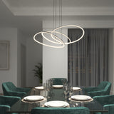 A modern dining room with a glass table set for six, surrounded by green velvet chairs. Two large, circular LED pendant lights hang above the table. A plant and large curtained window are in the background.