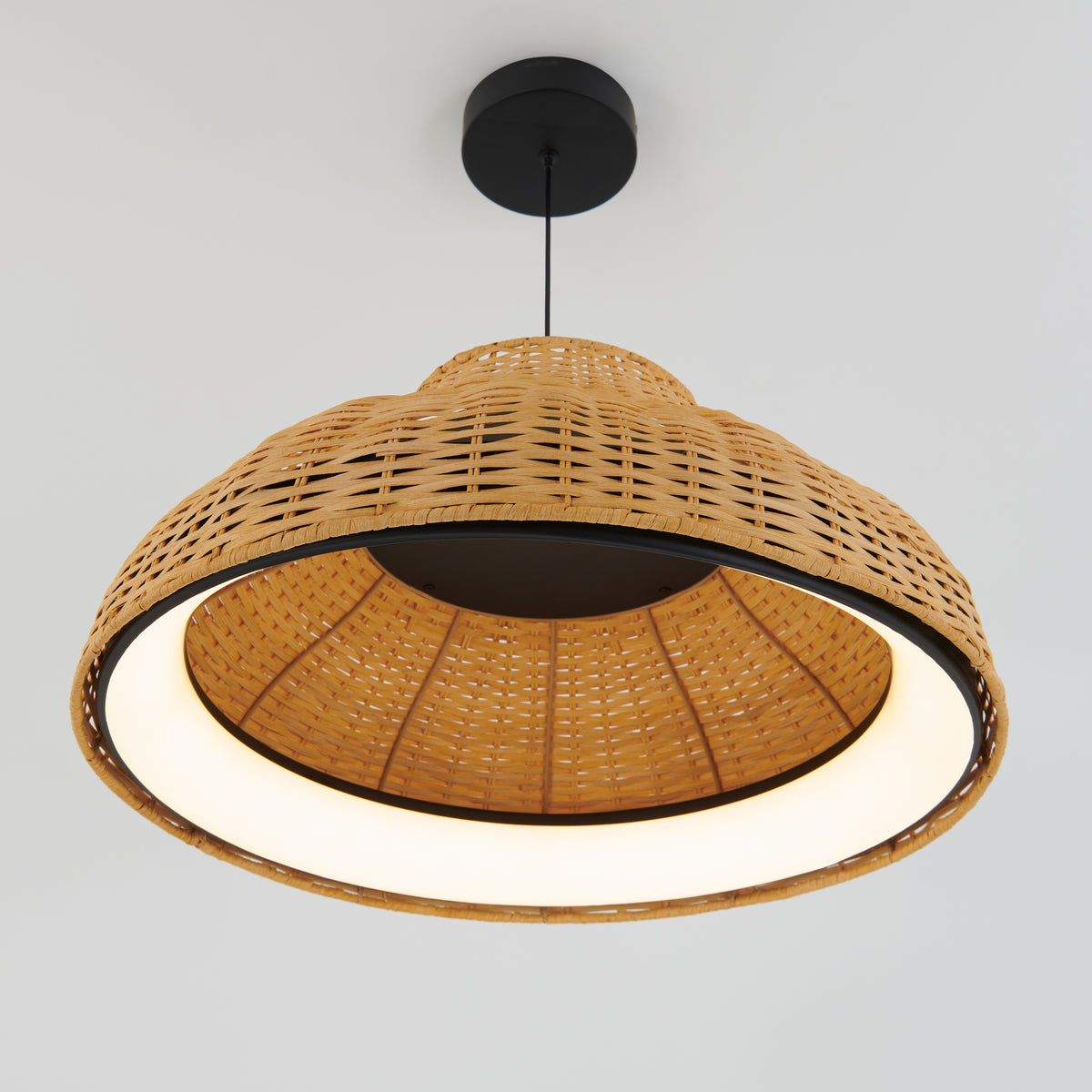 A woven rattan pendant light with a round, dome-shaped shade hanging from a black ceiling mount on a white background. The light is on, creating a warm, inviting glow inside the basket-like shade.
