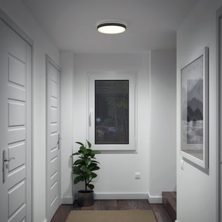 A modern, white hallway with three closed doors, a window at the end above a potted plant, a framed picture on the right wall, dark wooden flooring, and an Artika Sunraker 10 in. LED modern flush mount ceiling light.