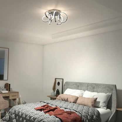 A modern bedroom with a grey upholstered bed, patterned bedding, and a red throw. An Artika Swirl Ribbon LED flush mount light shines overhead. In the corner, a desk with mirror, chair, and decor items completes the look.