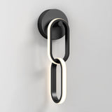Modern wall sconce with two interlocking oval rings, one illuminated, mounted on a round black base against a plain white wall. The design is minimalist and contemporary.
