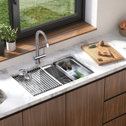 The Artika Newport double bowl stainless steel sink with a chrome faucet, built-in drying rack, and cutting board sits on a white marble countertop. Sliced figs, herbs, and kitchen tools are arranged by a window overlooking the garden.