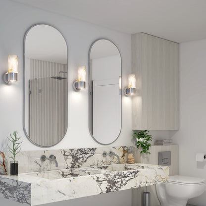 Modern bathroom featuring two oval mirrors, a marble countertop with dual sinks, Artika Marble LED contemporary sconces, silver fixtures, potted plants, light wood cabinetry, and a toilet with toilet paper on the right.