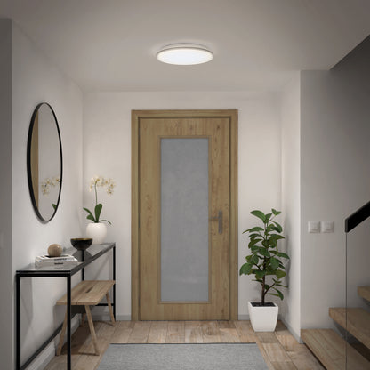 A modern entryway featuring a wooden door, black-framed console table, round mirror, wooden bench, potted orchid and plant, staircase with glass railing, and illuminated by the Artika Astrid 16.5 in. LED flush mount light (5 CCT) with remote.