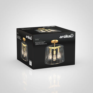 A black Artika box displays the Gatsby flush mount light, a modern gold and black ceiling fixture with exposed bulbs and a metal cage design. The packaging shows the brand name and product details.