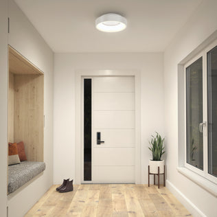 A modern entryway featuring wood flooring, a white door, a built-in bench with pillows, a potted plant on a stand, brown shoes on the floor, and bright illumination from the Artika Aiden LED flush mount ceiling light.