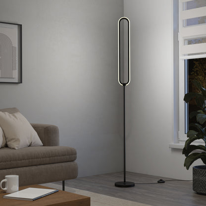 A modern living room features a beige sofa, coffee table, framed abstract print, and the Artika Leni dimmable LED modern floor lamp with an oval shape beside a window with blinds and a large green plant.