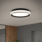 A modern, round LED ceiling light with a black frame and a decorative, textured band emits a soft, white glow in a minimalist room with gray and wooden walls.
