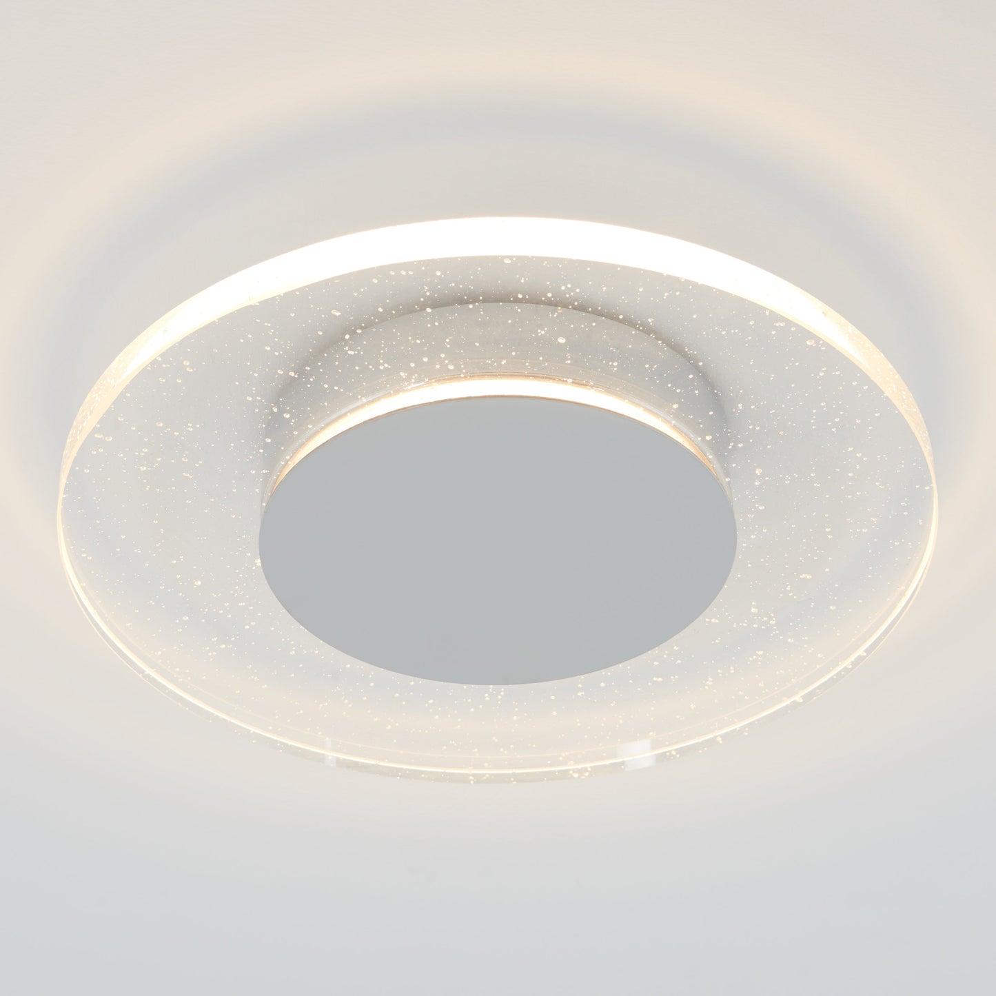 The Essence Disk LED flush mount by Artika-CAN-dev features a chrome finish, a central disc, and a clear ring with sparkling particles, emitting soft white light through an elegant glass diffuser.