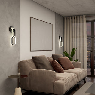 Modern living room featuring a beige sofa, brown and tan pillows, abstract art, Artika’s Catena modern LED wall sconce (5 CCT), a small round side table, potted plant, and sheer curtains over tall windows.