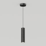 A modern black cylindrical pendant light hangs from the ceiling by a thin cord. The lampshade has small perforated holes, allowing light to shine through, and casts a subtle glow below against a plain white background.