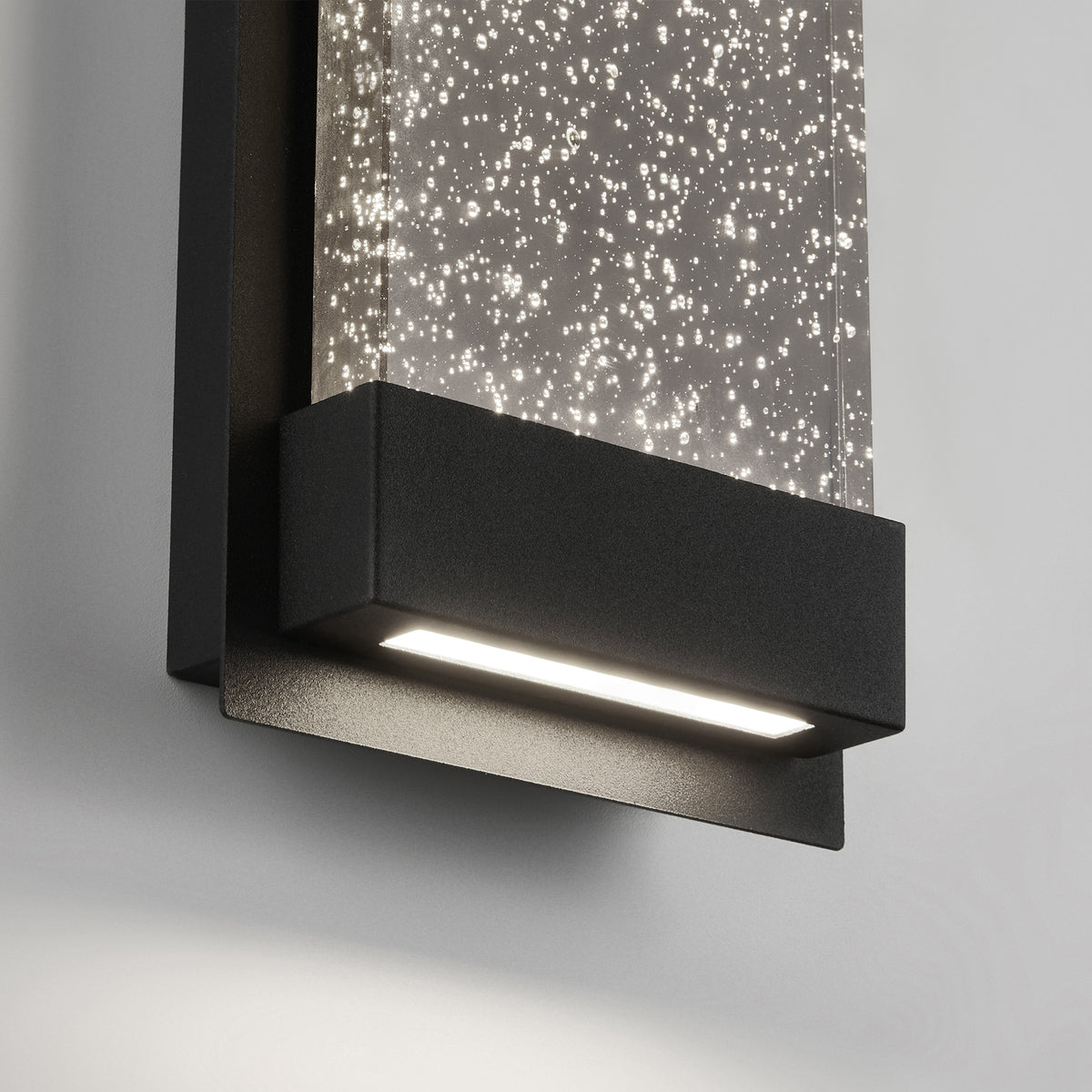 A modern wall-mounted light fixture with a black frame and a clear panel filled with small, sparkling bubbles, emitting a soft white light onto a light gray wall.