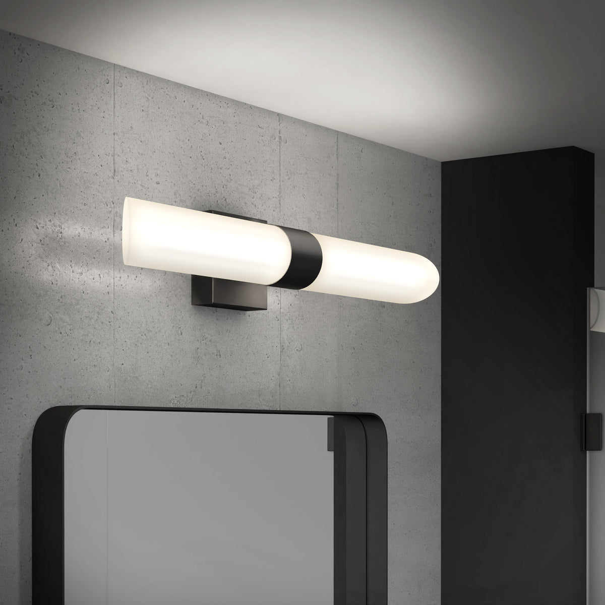 Modern wall-mounted cylindrical light fixture with a black base, installed above a large mirror on a gray concrete wall in a contemporary bathroom setting.