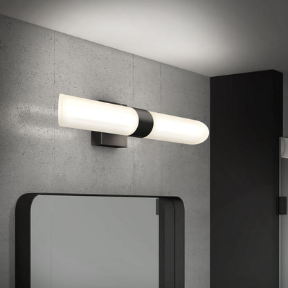 The Artika Wilton LED modern bathroom vanity light 5 CCT, featuring a cylindrical white shade with black accents, is mounted above a rectangular mirror on a gray concrete wall in a contemporary bathroom.
