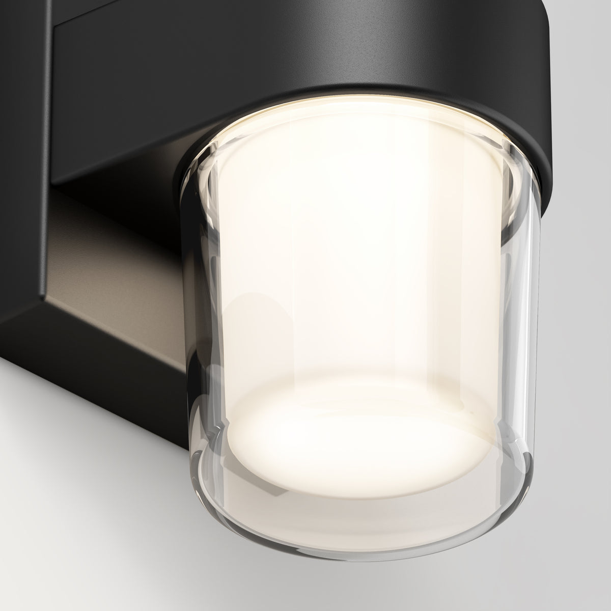 Close-up of a modern wall-mounted light fixture with a cylindrical glass shade and black metal base, illuminated and casting a soft, warm glow.