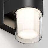 Close-up of a modern wall-mounted light fixture with a cylindrical glass shade and black metal base, illuminated and casting a soft, warm glow.