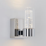 A modern wall sconce with a square chrome base and a cylindrical glass shade, featuring bubbles inside the glass, mounted on a light-colored wall.