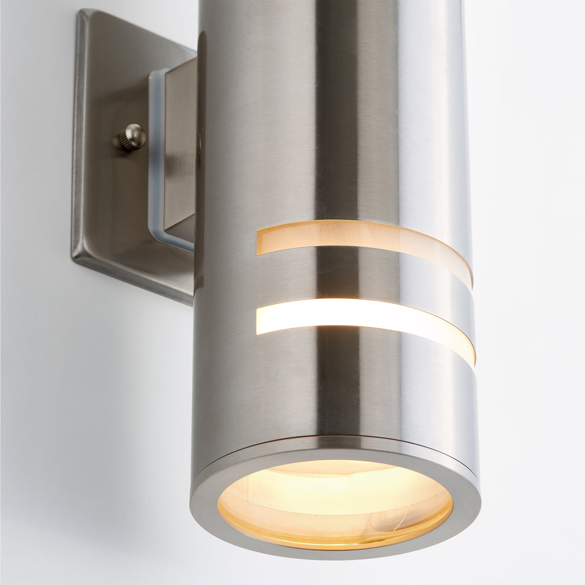 A modern wall-mounted cylindrical stainless steel light fixture with horizontal cut-out slits allowing light to shine through, and a frosted circular glass cover on the bottom, attached to a square wall plate.