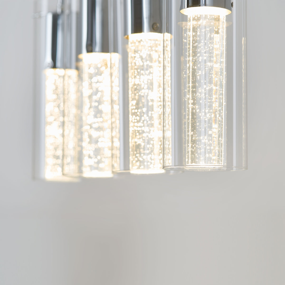 Close-up of the Artika Bubble Flow 4-light pendant, featuring modern cylindrical glass shades with bubbles inside, emitting a bright white glow against a plain light background.