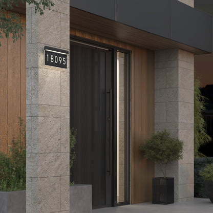Modern house entrance with a black door, stone and wood-paneled walls, two potted plants, and an illuminated address plaque displaying the number 18095.