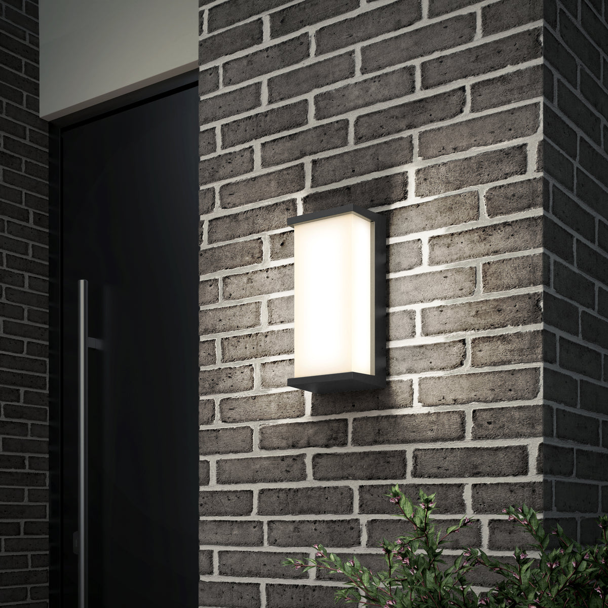A modern rectangular wall light with a frosted cover is mounted on a brick exterior wall next to a dark-colored door, with green foliage visible at the bottom right corner.