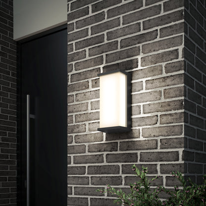 The Artika Faro modern LED indoor-outdoor wall light 3 CCT, featuring frosted glass and a rectangular design, is mounted on a gray brick wall near a doorway with a small green plant in the bottom right corner.