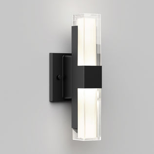 The Artika Gemini indoor-outdoor LED wall light features a rectangular black metal base and a vertical frosted glass fixture, casting a soft white glow against a light gray wall.