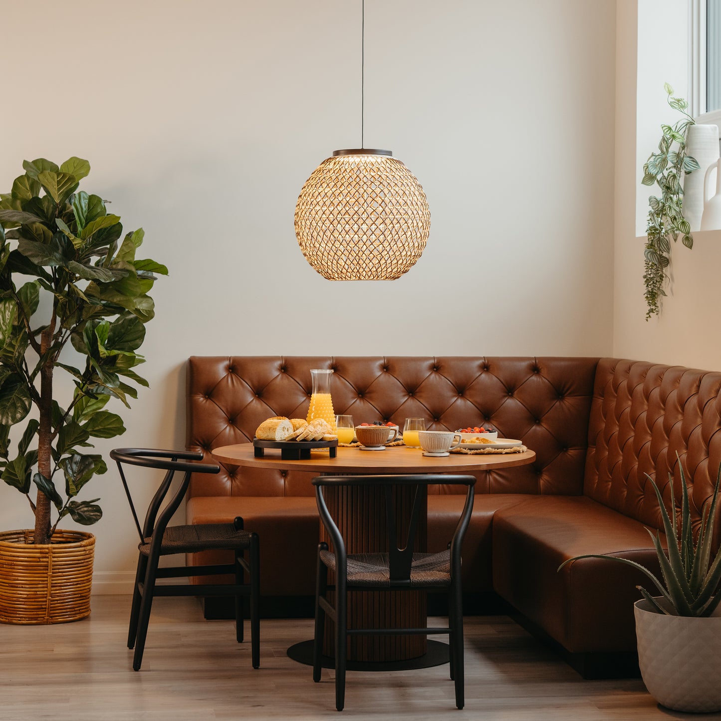 A cozy dining nook with a tufted brown leather bench, round wooden table set for breakfast, black chairs, a potted plant, window-hanging greenery, and the Artika Benson LED pendant light 5 CCT illuminating the space.