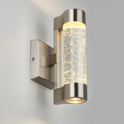 The Artika Bubble Element LED indoor-outdoor light is a modern, wall-mounted fixture with a brushed metal finish and cylindrical design. Its clear bubble section glows with warm light, shown here on a light gray wall.