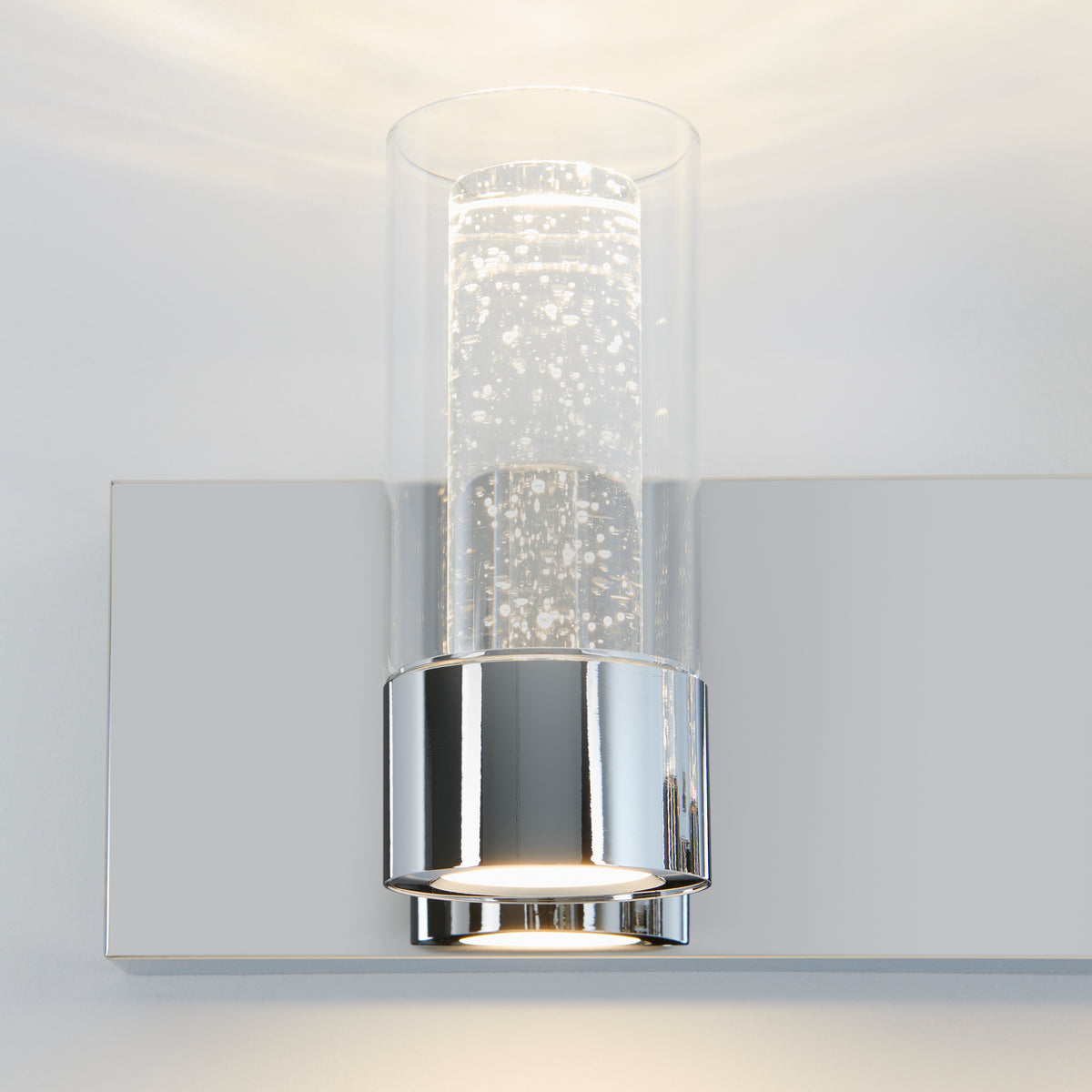 A modern wall sconce with a chrome base and a cylindrical glass shade featuring suspended air bubbles, mounted on a light gray wall and emitting soft white light.