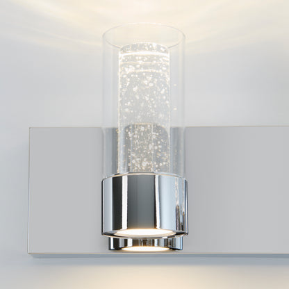 The Artika Ratio 2-light LED bathroom vanity light features a chrome base and cylindrical bubble glass shades, casting soft white light on a light-colored wall.