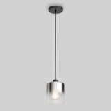 A modern pendant light with a cylindrical clear glass shade hangs from a black cord and ceiling mount against a plain light gray background. The bulb inside the shade is illuminated.