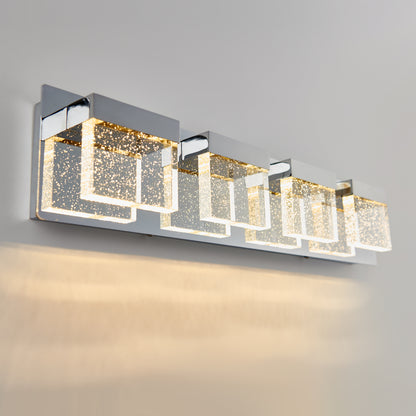 The Artika Bubble Cube 4-light LED bathroom vanity light features four rectangular glass shades filled with sparkling bubbles, mounted on a sleek chrome base, casting a warm glow against a light gray wall.