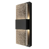 A modern wall sconce with a rectangular black base and a textured, frosted glass panel featuring speckled patterns. Soft, warm light glows from behind the panel, creating an ambient effect.