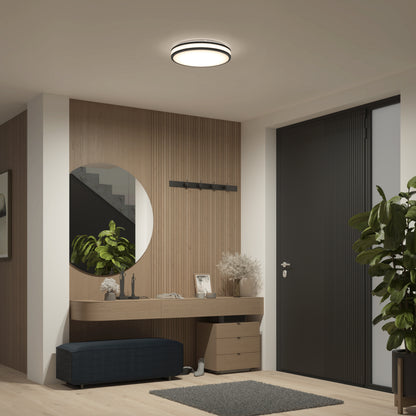 A modern entryway with wood paneling, a round mirror, wall-mounted hooks, a console table with decor, a small bench, and potted plants is beautifully lit by the Artika Hemisphere 15 in. LED flush mount light 5 CCT for adjustable brightness.