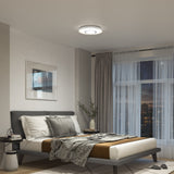 A modern bedroom with a low platform bed, neutral bedding, a side table with a lamp, a large window with sheer curtains, a framed picture on the wall, and a round ceiling light.
