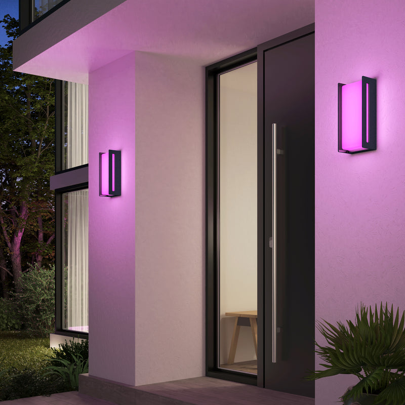 A modern entrance features two Artika Ryker smart RGB indoor-outdoor wall lights glowing pink on white walls beside a glass door. A bench is visible inside, with plants in the foreground.