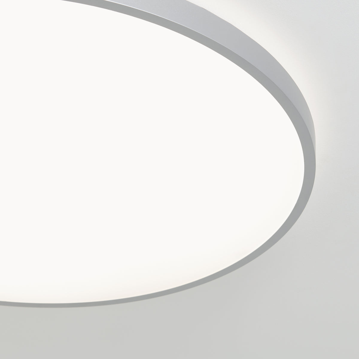 A close-up view of a round ceiling light fixture with a smooth white surface and a thin gray rim, glowing brightly against a plain white ceiling.