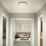 A modern hallway leads to a living room with a gray sofa, orange blanket, two abstract wall art pieces, and a round coffee table with flowers and books. Light wood flooring runs throughout the space.