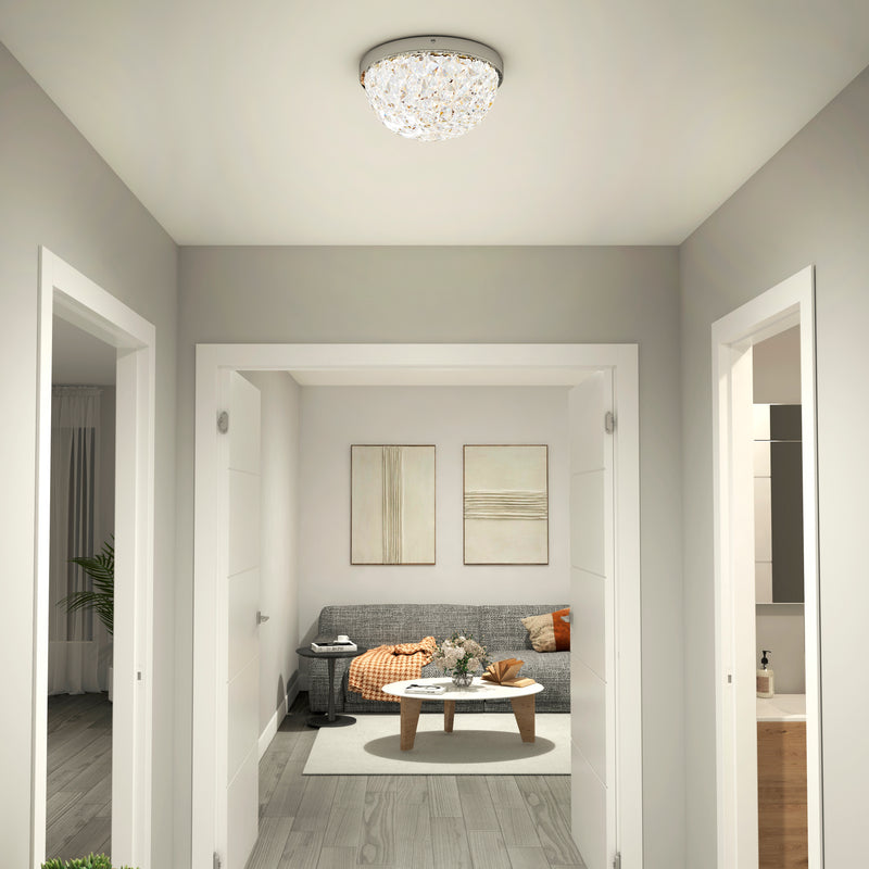 A modern hallway with light gray walls and wood floors leads to a cozy living room with a gray sofa, orange blanket, coffee table, and abstract wall art. The space is illuminated by the Artika Carat LED flush mount light.