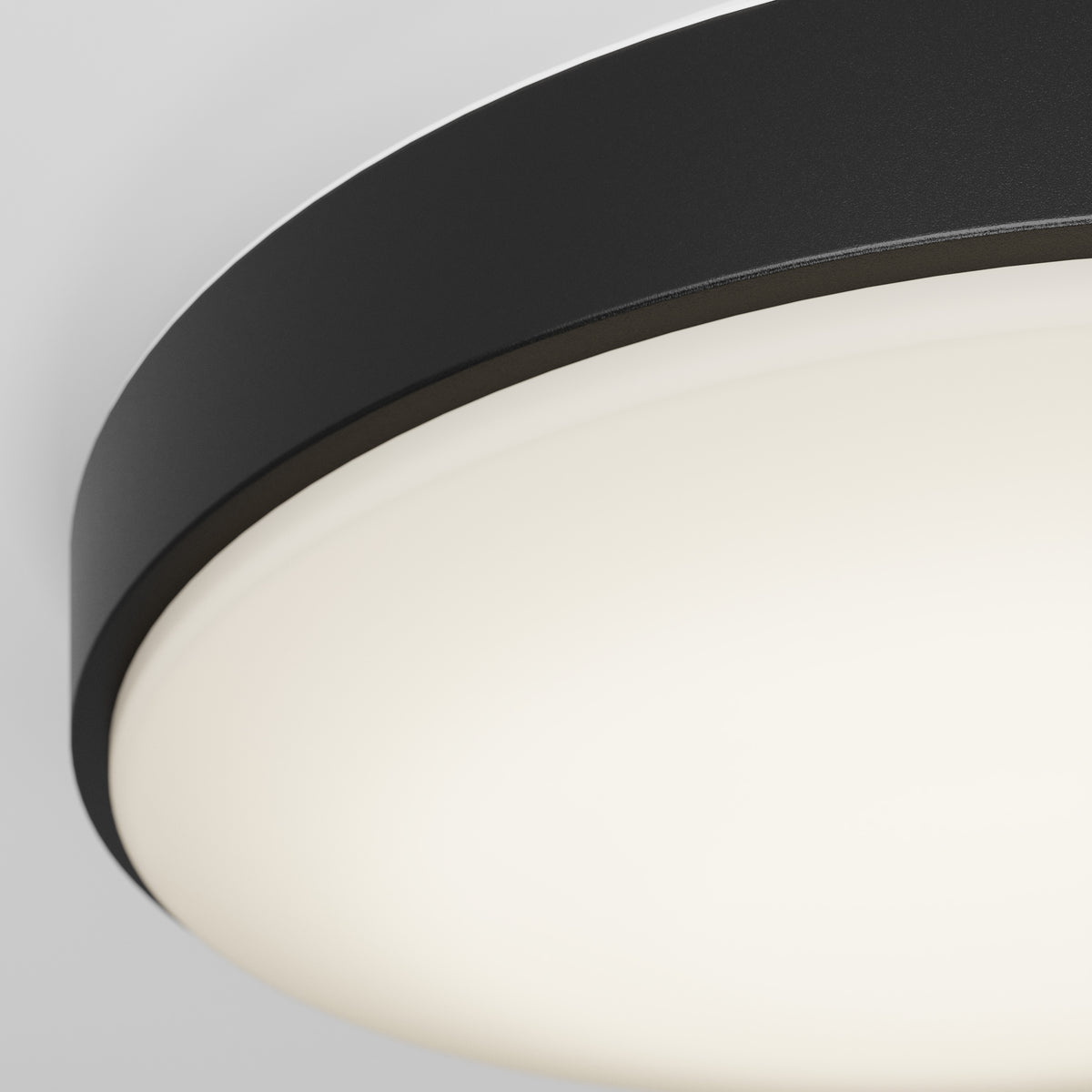 Close-up of a modern, round ceiling light fixture with a black rim and a white, glowing diffuser against a light gray background.
