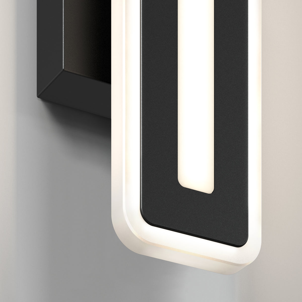 Close-up of a modern wall light fixture with a rectangular black frame and rounded corners, featuring a glowing white LED strip outlining its shape, mounted on a light-colored wall.