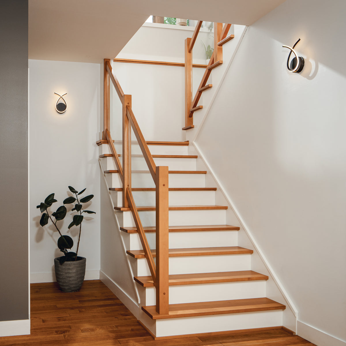 Wooden staircase with glass railing, white walls, Artika Gamma modern LED wall sconces, hardwood floor, and a potted plant.