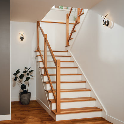 Wooden staircase with glass railing, white walls, Artika Gamma modern LED wall sconces, hardwood floor, and a potted plant.
