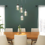 A modern dining room with a wooden table, six beige upholstered chairs, a small glass vase with green leaves, and four glass pendant lights hanging above. Two large windows with sheer white curtains let in natural light.