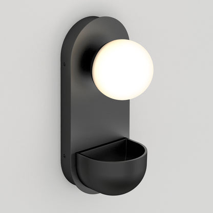 The Artika Bloomfield LED modern mid-century wall sconce 5 CCT features a matte black finish, a round frosted glass globe on top, and a semi-circular storage compartment, shown mounted on a pale gray wall.