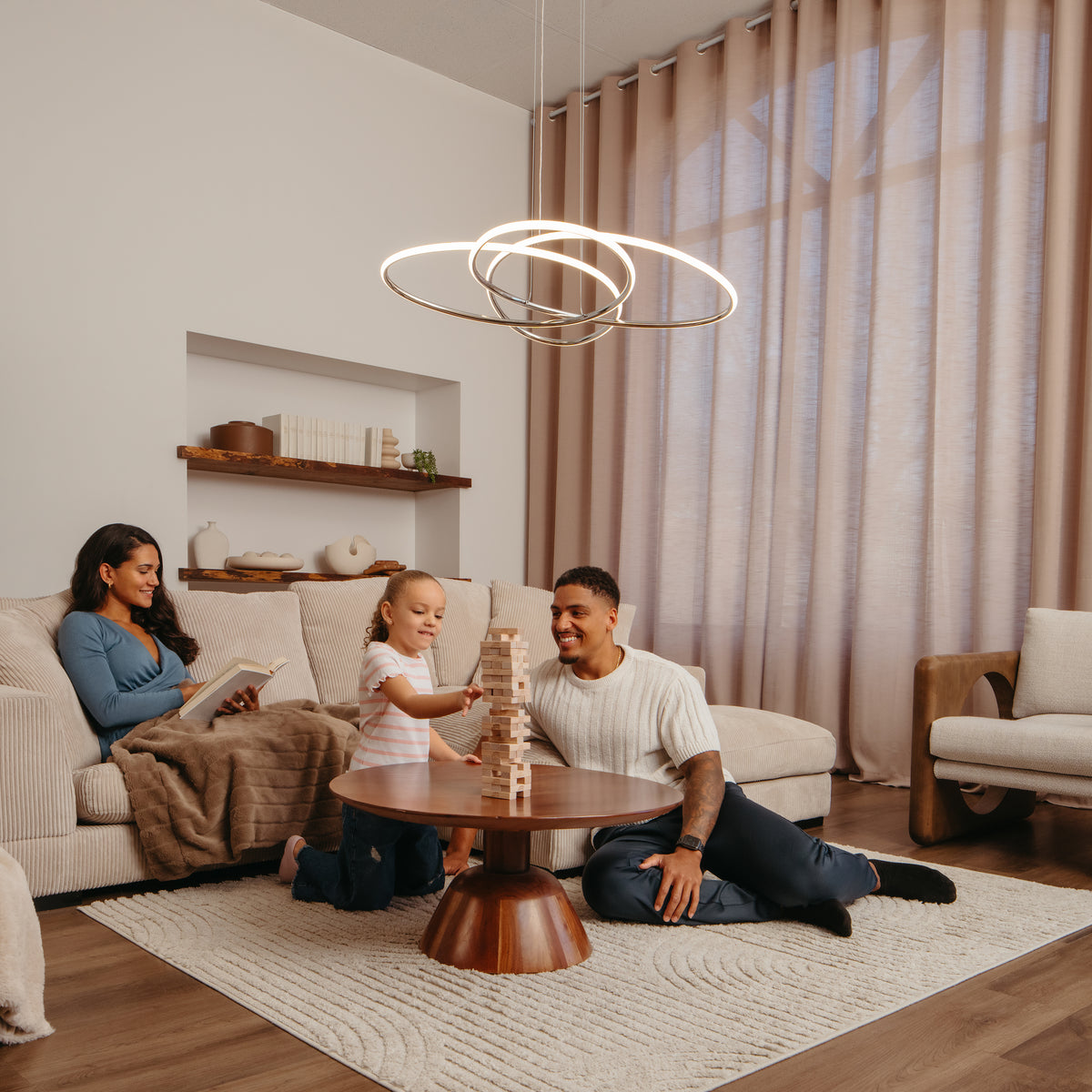 A family relaxes in a cozy living room; a woman sits on a couch reading, while a man and young girl play a block-stacking game on the floor around a wooden coffee table.