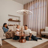 A family relaxes in a cozy living room; a woman sits on a couch reading, while a man and young girl play a block-stacking game on the floor around a wooden coffee table.