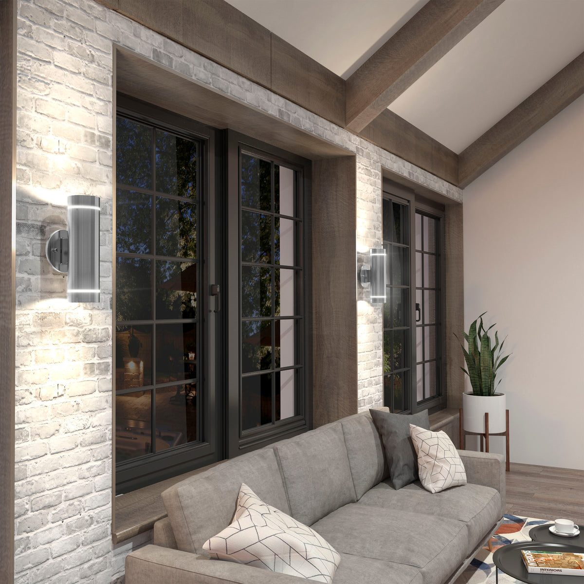 A modern living room with large windows, exposed brick walls, Artika C7 indoor-outdoor wall lights, a gray sectional sofa with patterned pillows, a plant on a stand, and a coffee table with magazines.